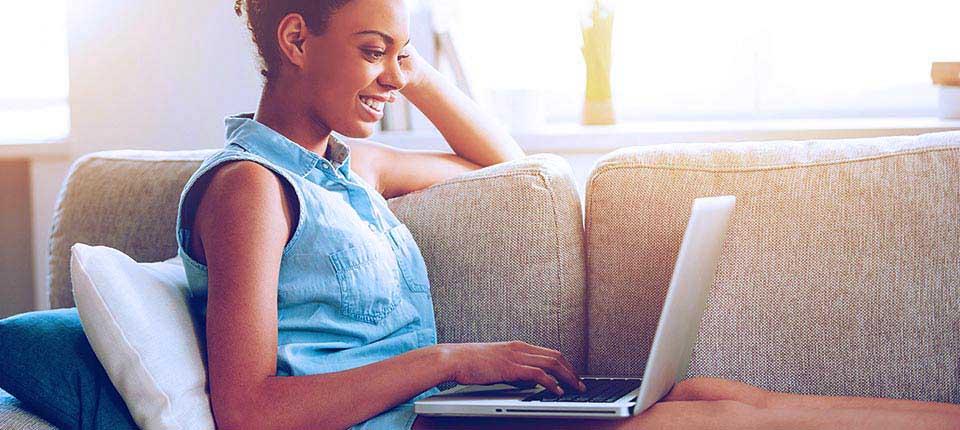 Surfing web at home. Side view of attractive young African woman working on laptop and smiling while sitting on the couch at home