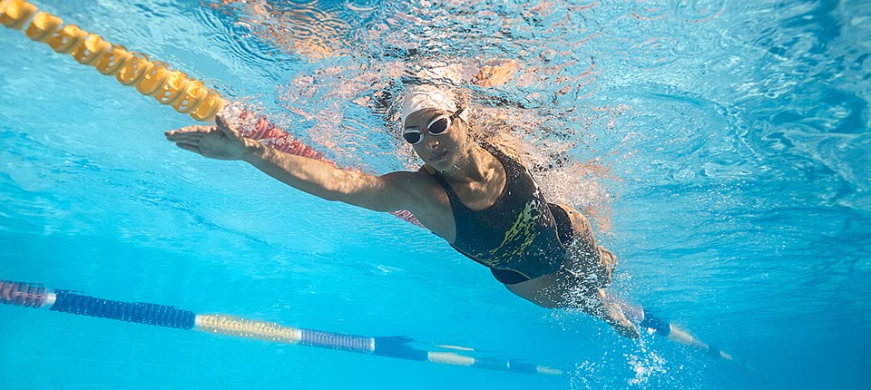 Beautiful girl swims underwater in the swimming pool outdoors. She wears a black-gray swimsuit with patterns, white swim cap and swim glasses. Sunlight falls from above. Splashes are around her body.