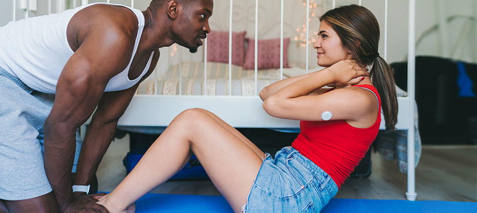 Young man helping his girlfriend to exercise regularly at home for healthy management of diabetes. She is wearing small sensor on the back of upper arm for measuring glucose levels and checking them on smartphone after scanning the sensor.