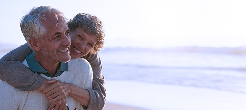 Portrait of happy mature man being embraced by his wife at the beach. Senior couple having fun at the sea shore.